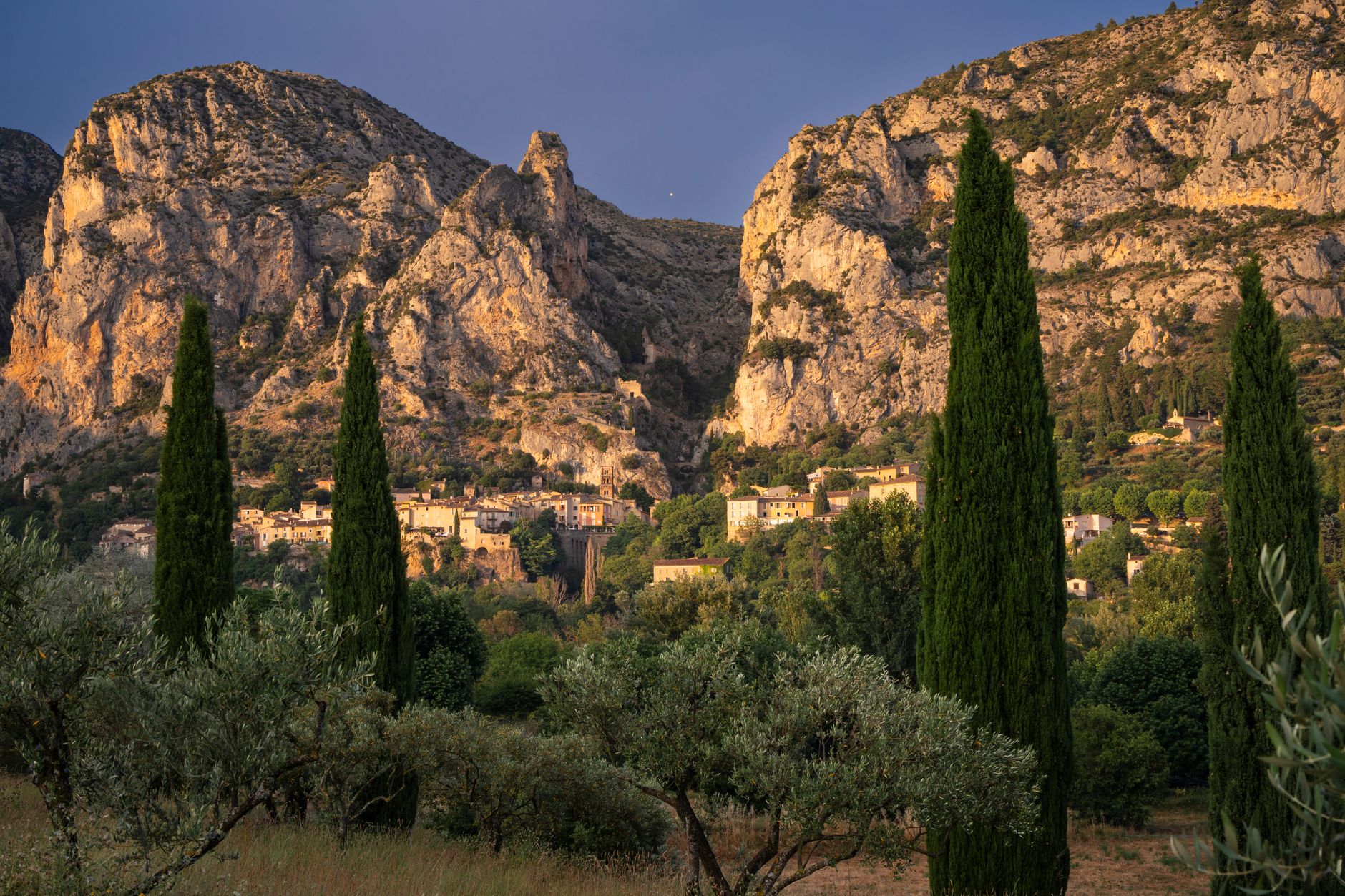 A scenic view of Moustiers-Sainte-Marie village with cypress trees and cliffs during golden hour in Provence, France. (Pexels, Le sixième rêve)